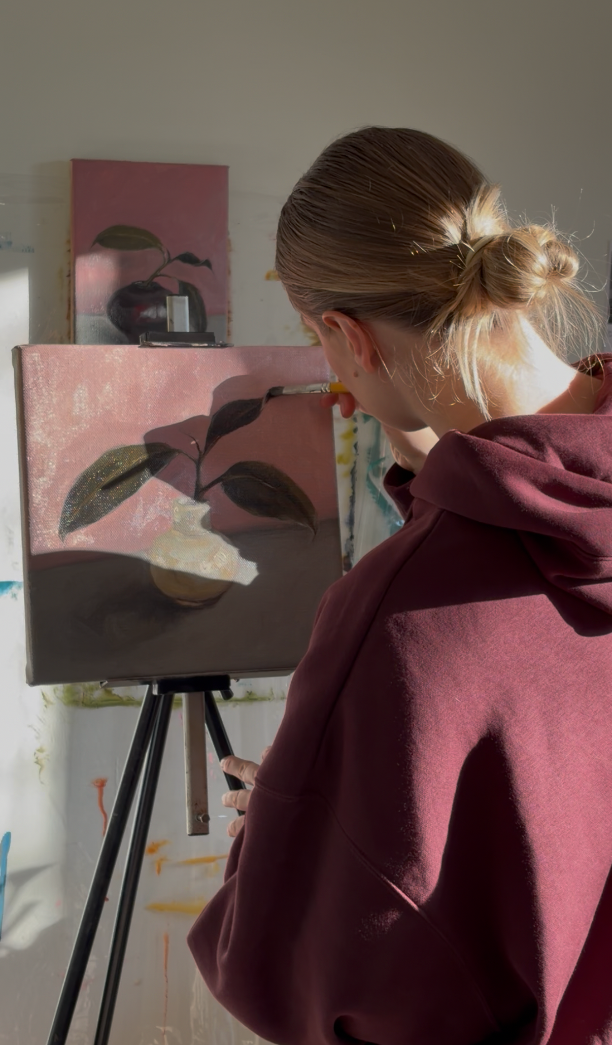 Person painting a still life scene on an easel in a room with light-colored walls.